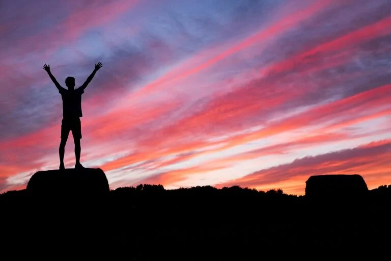 Silhouette of a person with arms raised on a clifftop under a pink sunset