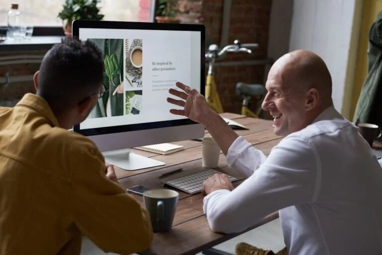Two colleagues smiling at a desktop monitor, one gesturing at the screen