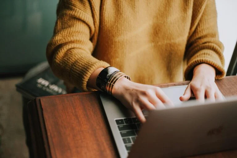 Person in a mustard sweater typing on a laptop at a wooden desk