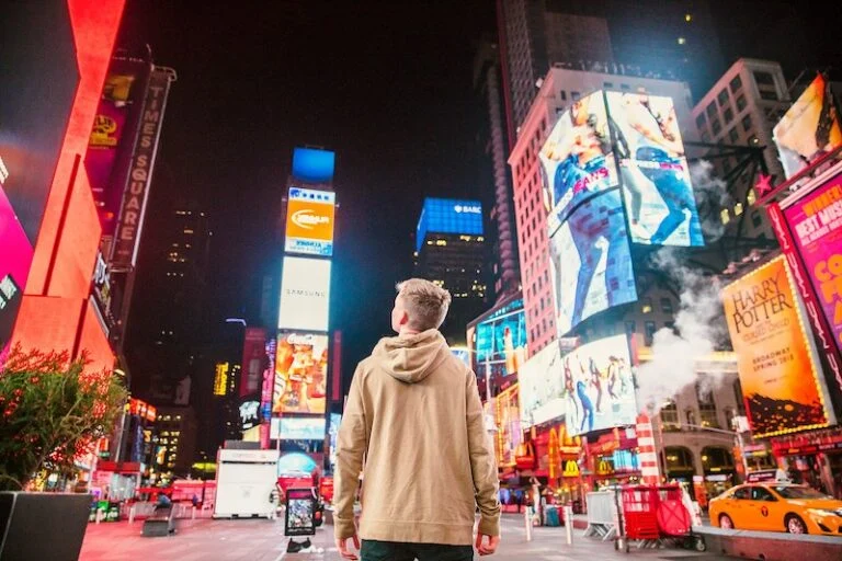 A person in a hooded sweatshirt standing in Times Square at night, surrounded by illuminated billboards