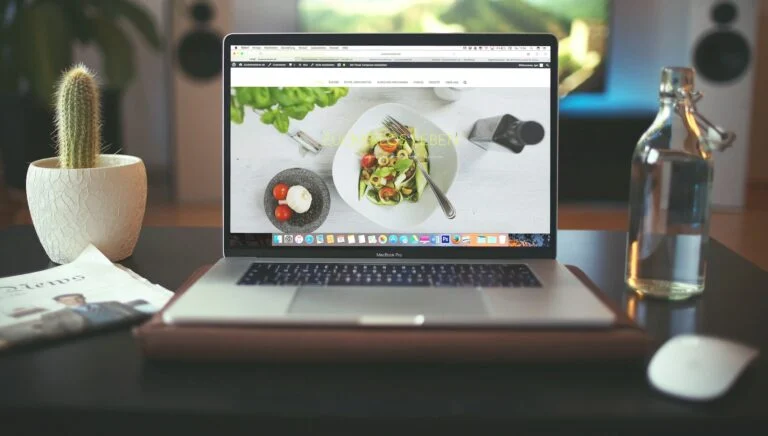 Laptop on a desk showing a plated dish, beside a small cactus and a bottle