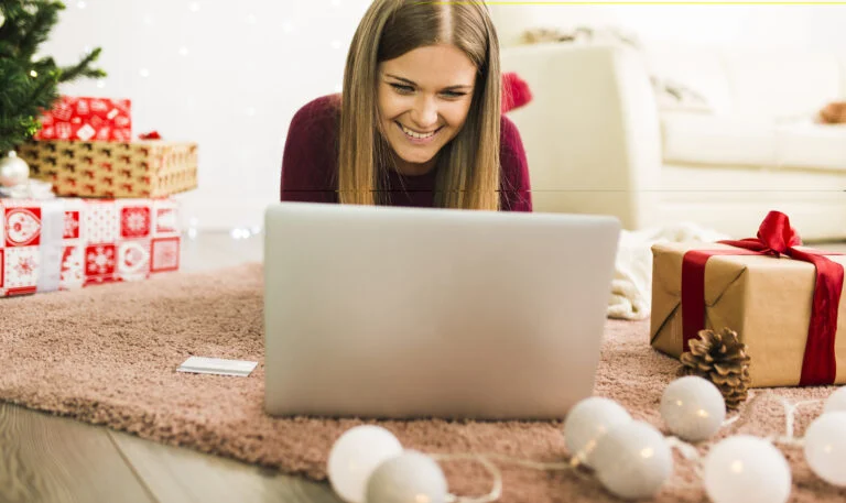 Woman smiling at a laptop beside wrapped holiday gifts