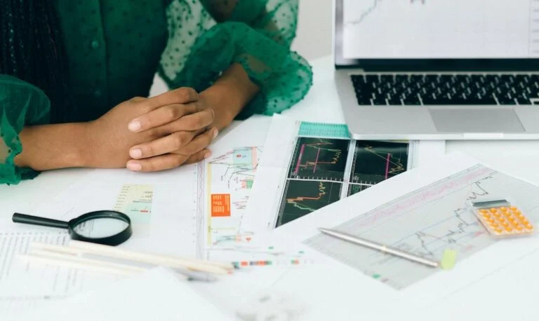 Hands resting on a desk spread with printed charts, a magnifying glass and a laptop